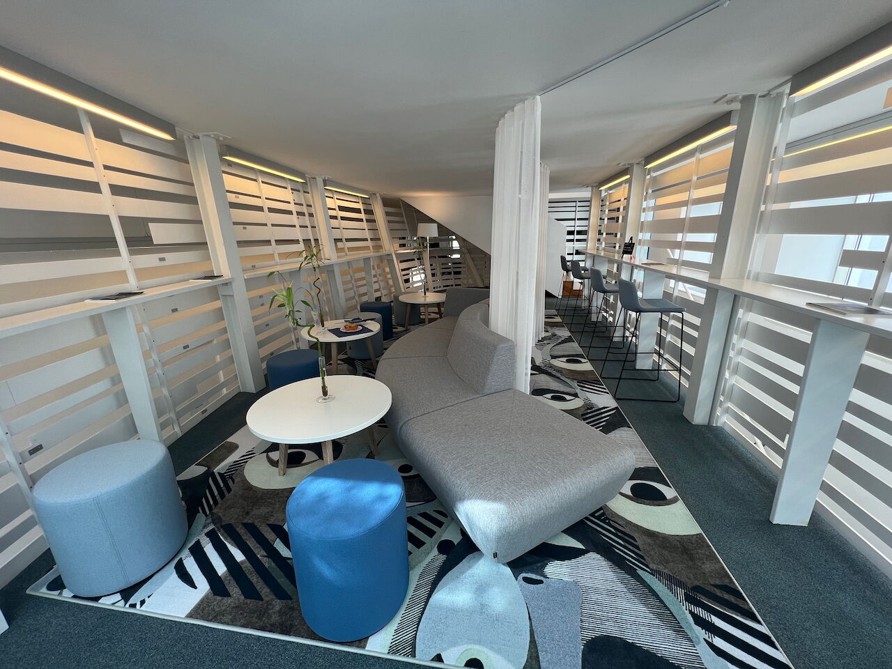 Spacious and modern office lounge interior featuring a large curved grey sofa, blue ottomans, white round coffee tables, and artistic patterned carpet.