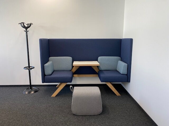 Blue high-back office booth seating with integrated wooden table, light blue cushions, grey stool, and coat rack in a modern office interior