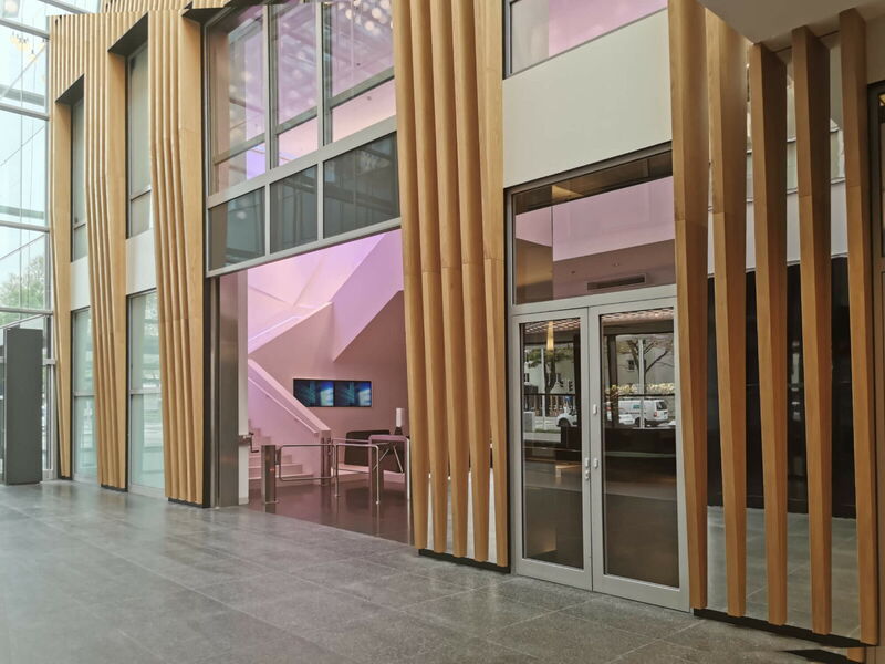 Interior view of a modern office lobby featuring vertical wooden slats, glass entrance doors, and a staircase with pink ambient lighting.
