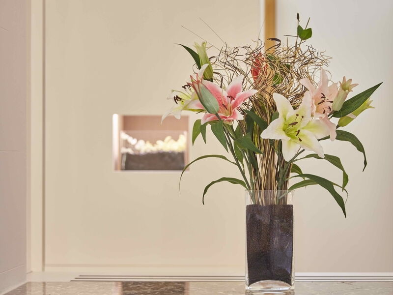 Wunderschöner Blumenstrauß aus rosa und weißen Lilien in einer rechteckigen Glasvase mit dekorativen Zweigen vor einer hellen Wand mit integrierter Beleuchtung.