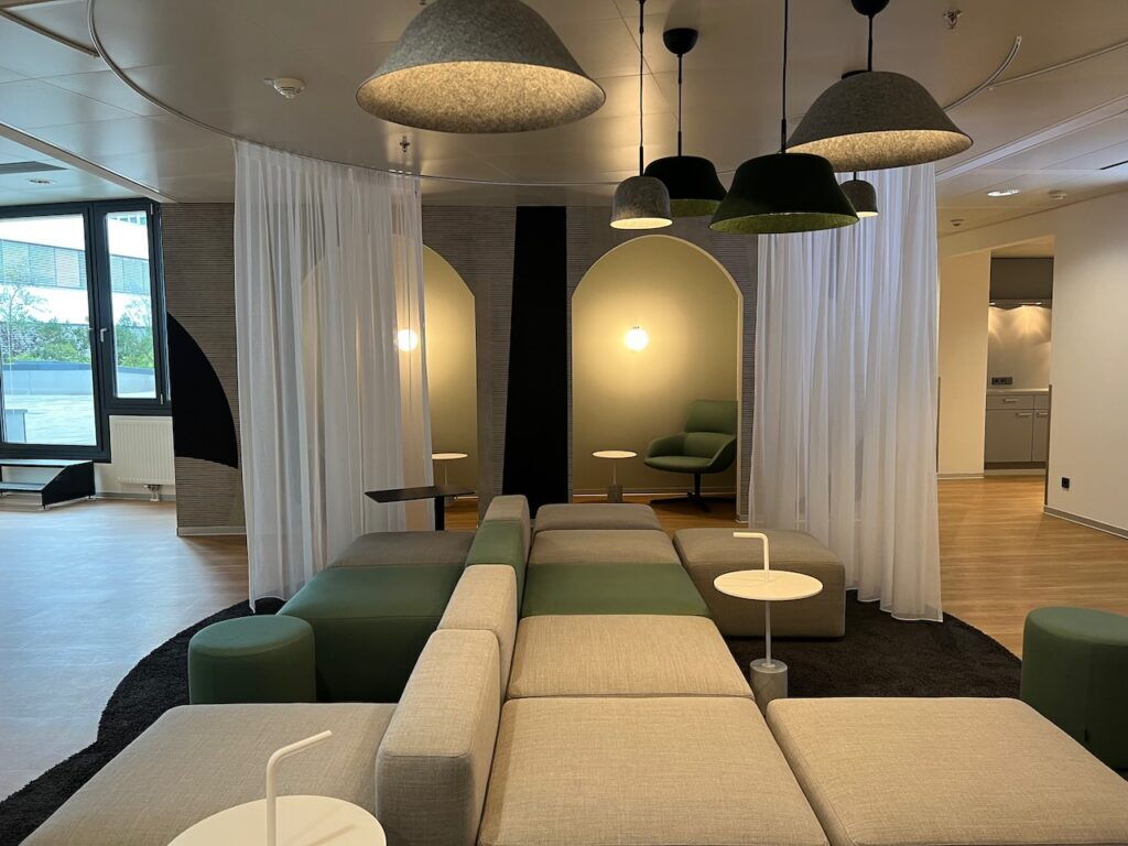 Spacious modern office lounge featuring modular green and grey seating, white sheer curtains, and stylish felt pendant lamps.