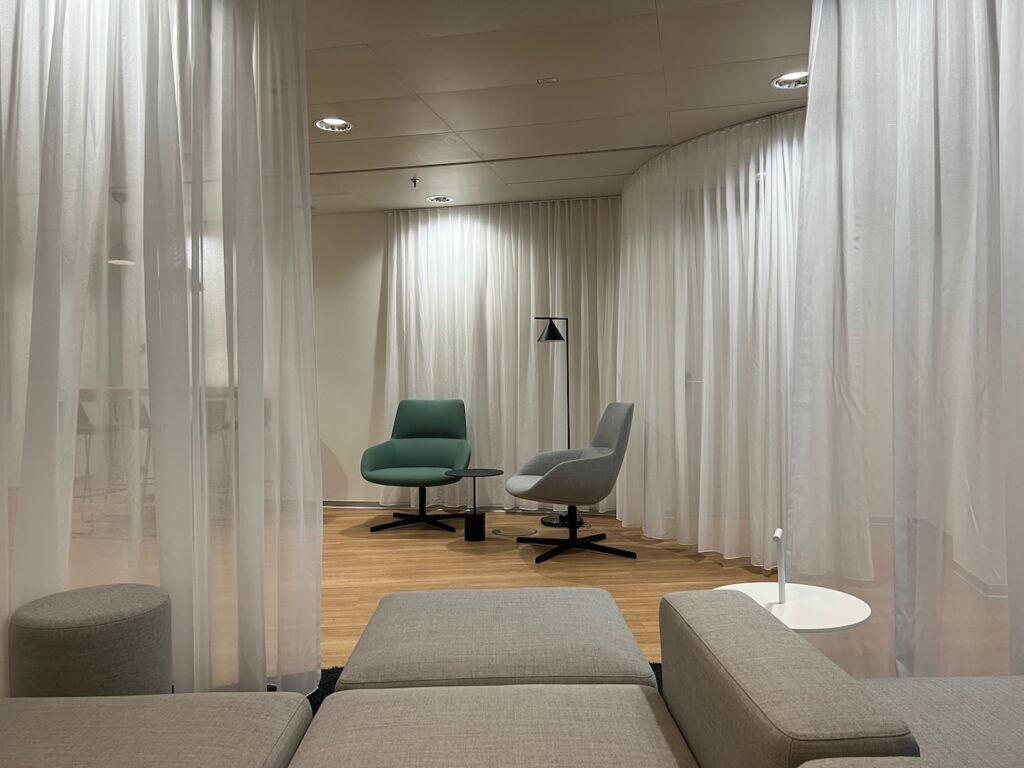 Modern office lounge space featuring white sheer curtains, a green armchair, a grey swivel chair, and comfortable seating.