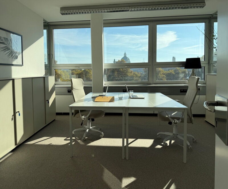 Heller moderner Büroarbeitsplatz im Akademiehof mit zwei Schreibtischen, ergonomischen Stühlen und großer Fensterfront mit Blick auf die Stadt