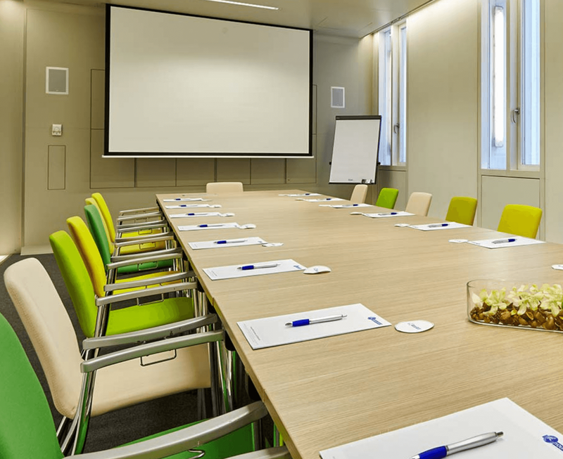 Bright conference room featuring a large projector screen, a long wooden meeting table with notepads and pens, and colorful chairs.