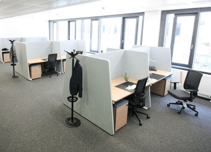 Overview of a modern office space featuring multiple cubicle desks with acoustic dividers, ergonomic chairs, and large windows.