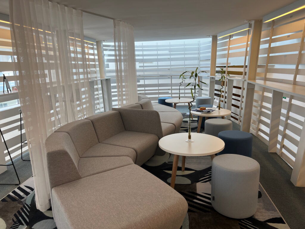 Modern office interior featuring curved grey sectional sofas, round coffee tables, and colorful stools in a bright work from anywhere lounge area.