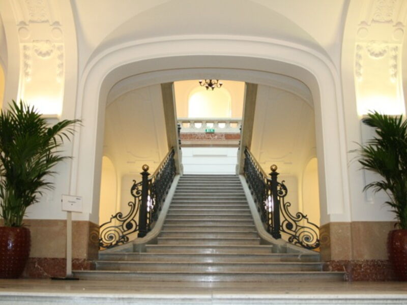 Eine prunkvolle Treppe mit verschnörktem schwarzem Eisengeländer in einem historischen Gebäude oder einer Hotelhalle, flankiert von großen Topfpflanzen und eleganten weißen Bögen.