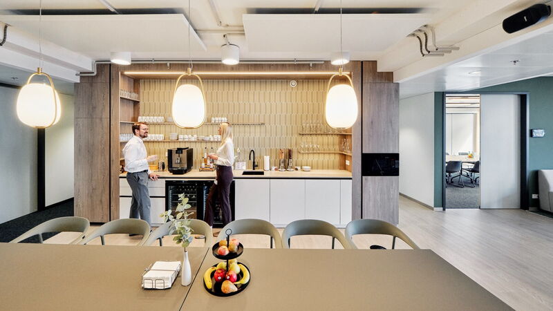 Two employees having a conversation in a modern, stylish office kitchen with coffee machine, wooden accents, and a large dining table in the foreground.