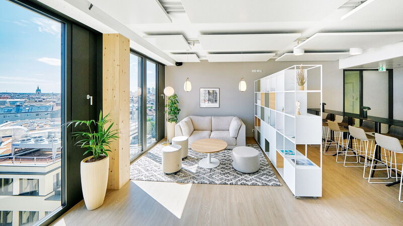 Bright and modern office lounge area featuring a comfortable sofa, coffee table, white shelving unit, and large windows with a city view.