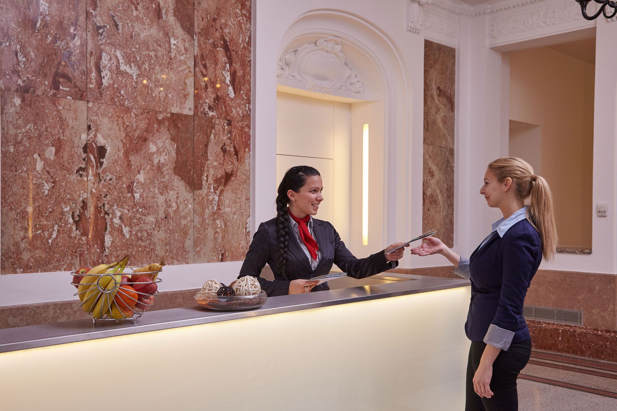 Eine lächelnde Rezeptionistin überreicht Dokumente an einen weiblichen Gast an einer eleganten Hotelrezeption