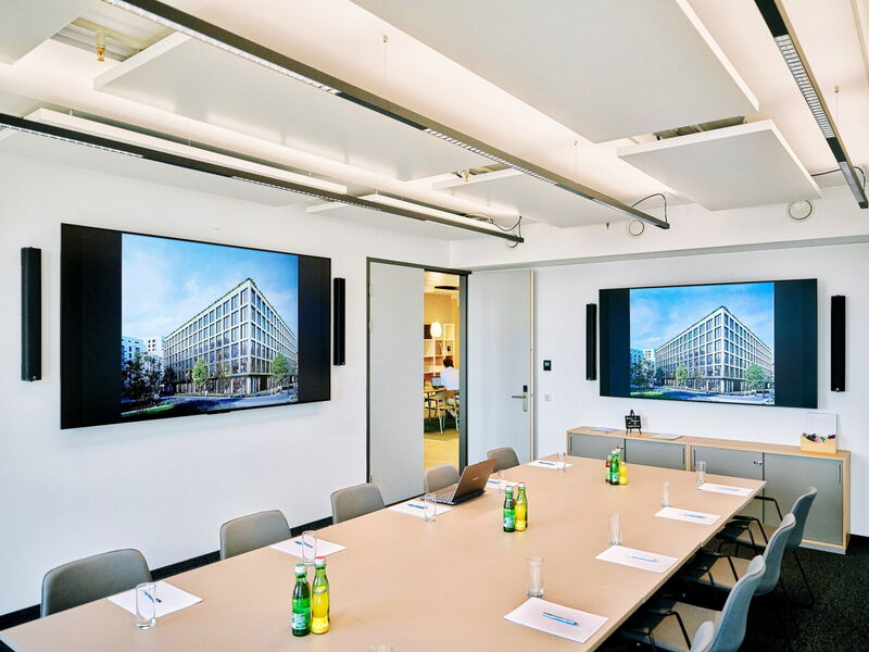 Modern conference room featuring a large wooden meeting table, ergonomic chairs, water bottles, and two large wall-mounted screens displaying architectural renderings.