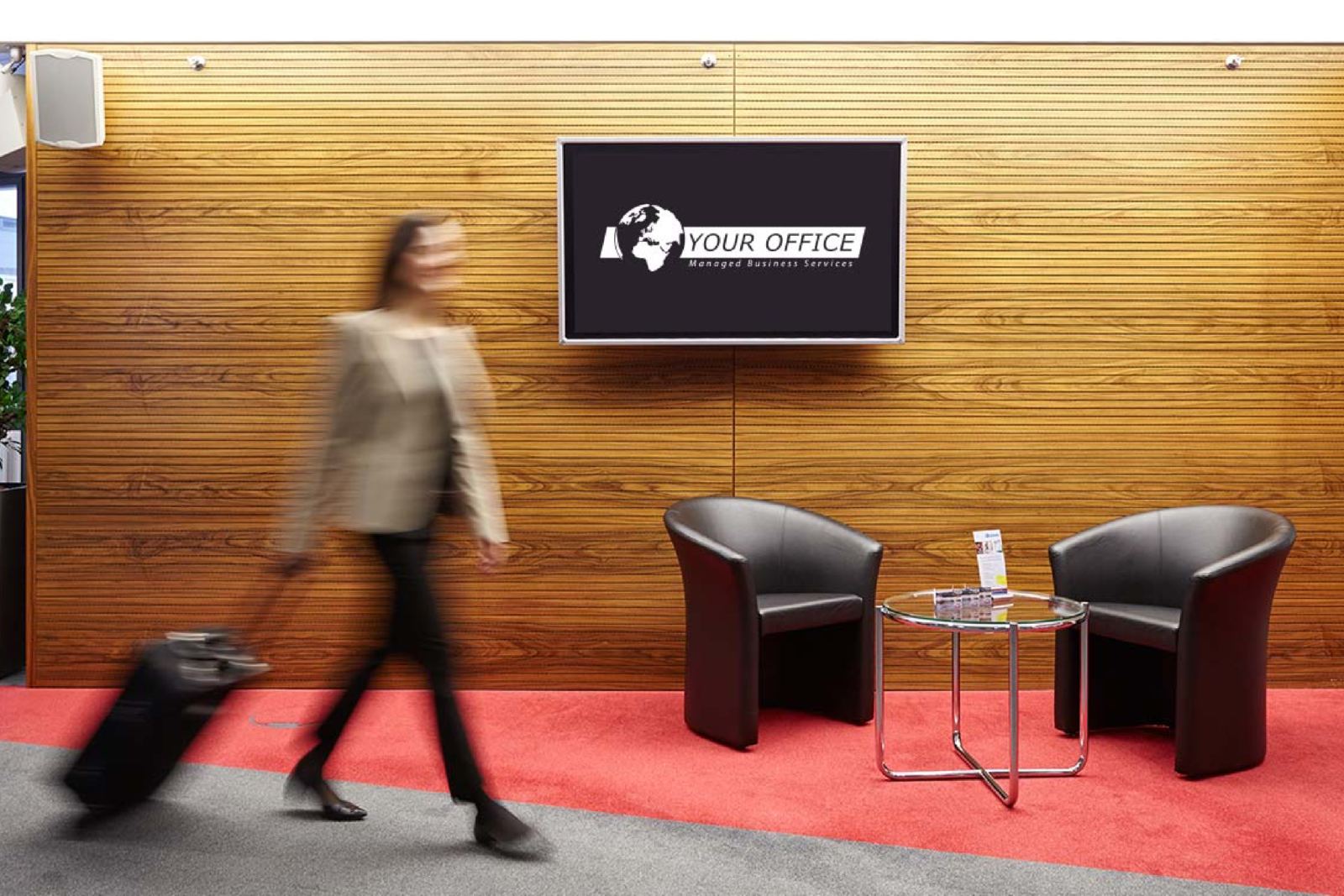 Eine Frau zieht einen Koffer durch ein modernes Bürofoyer mit digitaler Firmenlogo-Anzeige an der Holzvertäfelung, zwei schwarzen Sesseln und einem Glastisch.