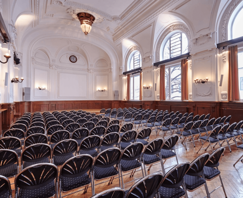 Geräumiger und eleganter Konferenzsaal mit klassischer Architektur, hohen Bogenfenstern und ordentlich angeordneten Sitzplätzen.