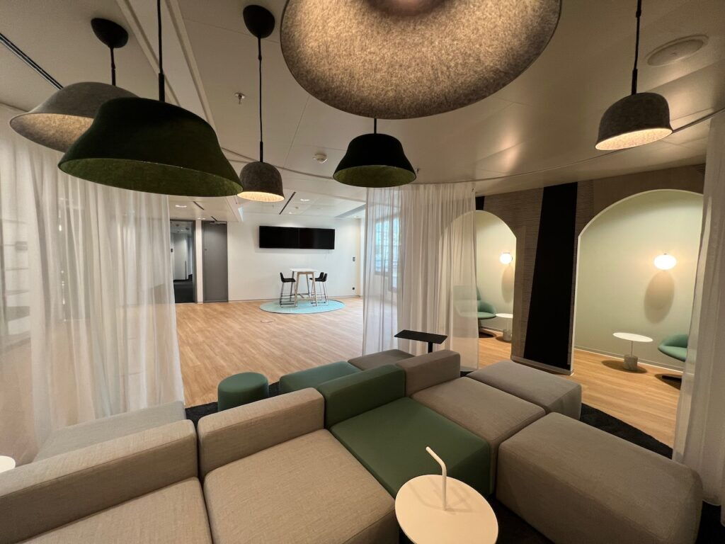 Spacious modern office lounge featuring modular grey and green sofas, unique felt pendant lamps, and private alcoves with white curtains.