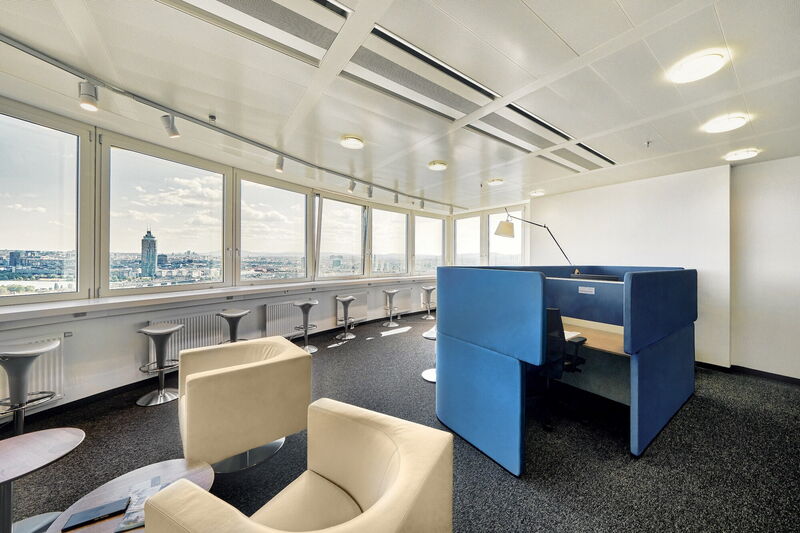 Modernes Büro mit einem blauen Akustik-Arbeitsplatz, cremefarbenen Sesseln, hohen Barhockern an großen Fenstern mit Panoramablick auf die Stadt und heller Beleuchtung.