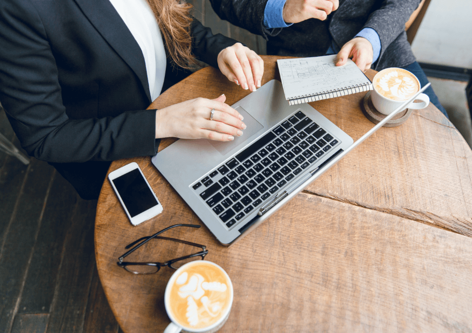 Man and woman in business attire working at a wooden table in a cafe with laptops, notepads, and coffee cups.