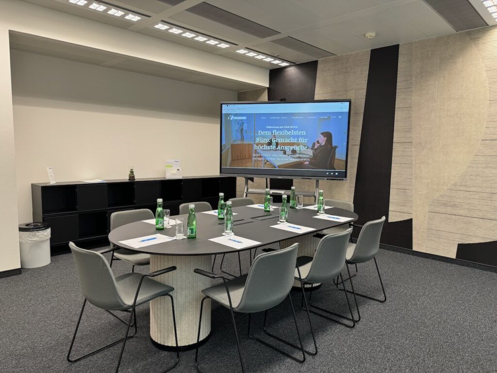 Bright modern meeting room equipped with a large oval table, comfortable grey chairs, and a presentation display screen for hybrid work and collaboration
