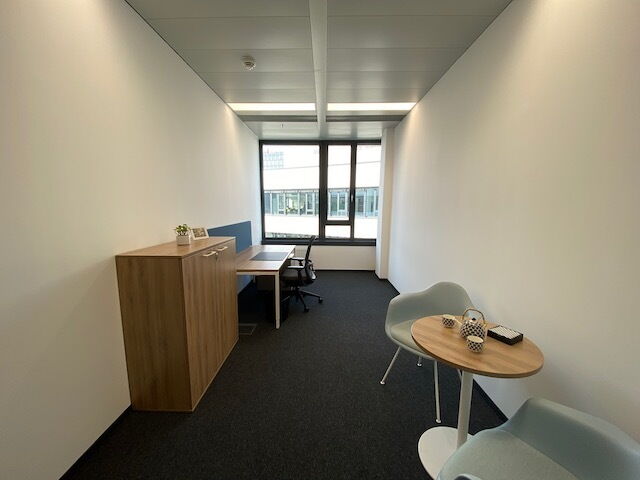 Ein helles, modernes Büro mit einem kleinen Besprechungstisch, hellblauen Sesseln, einem Holzschrank und einem Schreibtisch am Fenster.
